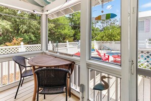Eine überdachte Terrasse im Freien mit einem Holztisch und Korbstühlen, einem weißen Zaun und Blick auf einen Garten mit Pool und roten Stühlen.