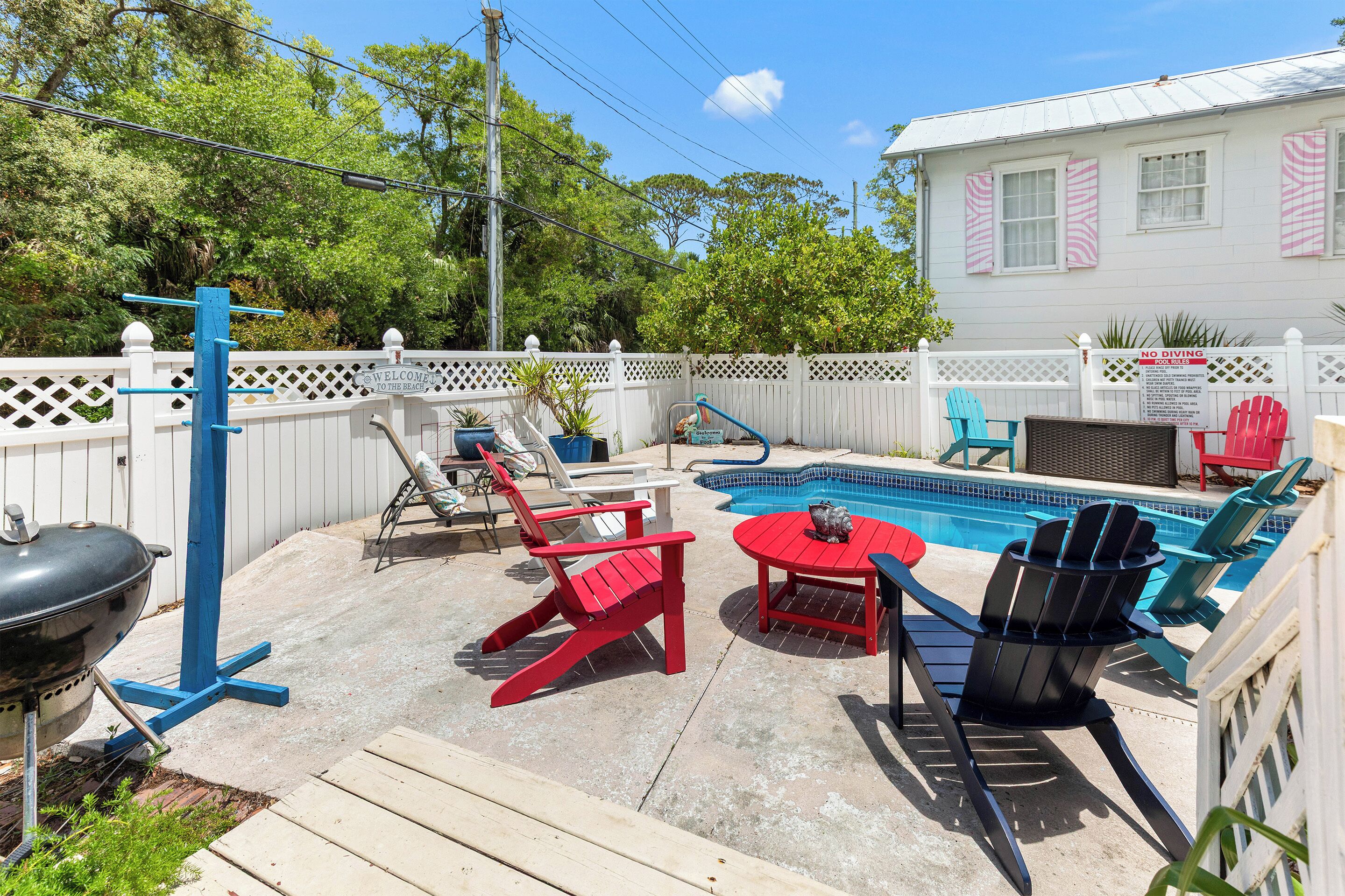 Un jardin avec une piscine, des fauteuils Adirondack rouges et noirs, un barbecue et une clôture blanche.