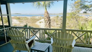 A covered outdoor area with two Adirondack chairs and a small table, overlooking a body of water and a sandy beach with vegetation. - Large, Classic Beachfront House with Wrap-Around Porch by Tybee Cottages (Tybee Island)