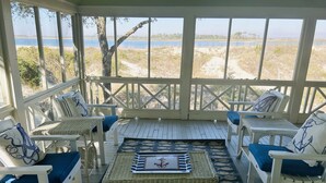 A covered outdoor seating area with wicker furniture and cushions, a table with a decorative tray, and a view of a beach and water. - Large, Classic Beachfront House with Wrap-Around Porch by Tybee Cottages (Tybee Island)