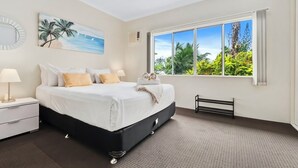 Un dormitorio con una cama grande, una mesita de noche, un ventilador de techo, una ventana que ofrece vistas a palmeras y a la playa, y un cuadro que representa una escena costera.
