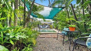 Un jardín tropical con piscina, rodeado de exuberante vegetación y una zona con sombra.