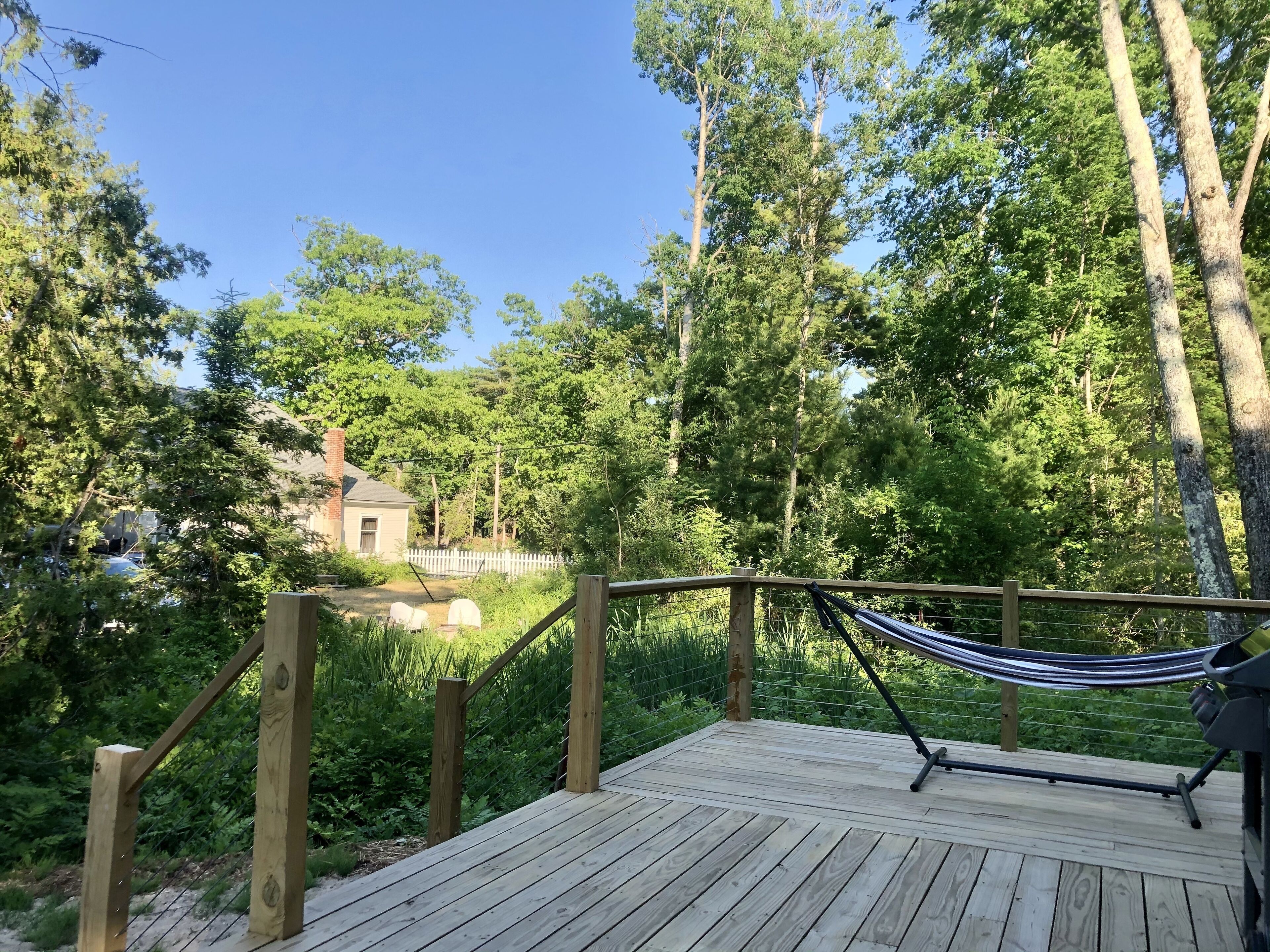 Une terrasse en bois avec un hamac, entourée d’une végétation luxuriante et d’une clôture blanche en bois.