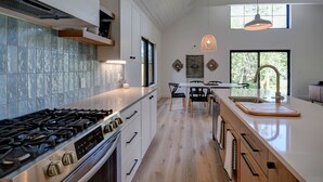 A modern kitchen with a stainless steel stove, white cabinetry, and a large island with a sink. - Modern home in Glen Arbor. Steam shower. Walk to lake. Air conditioning. (Glen Arbor)