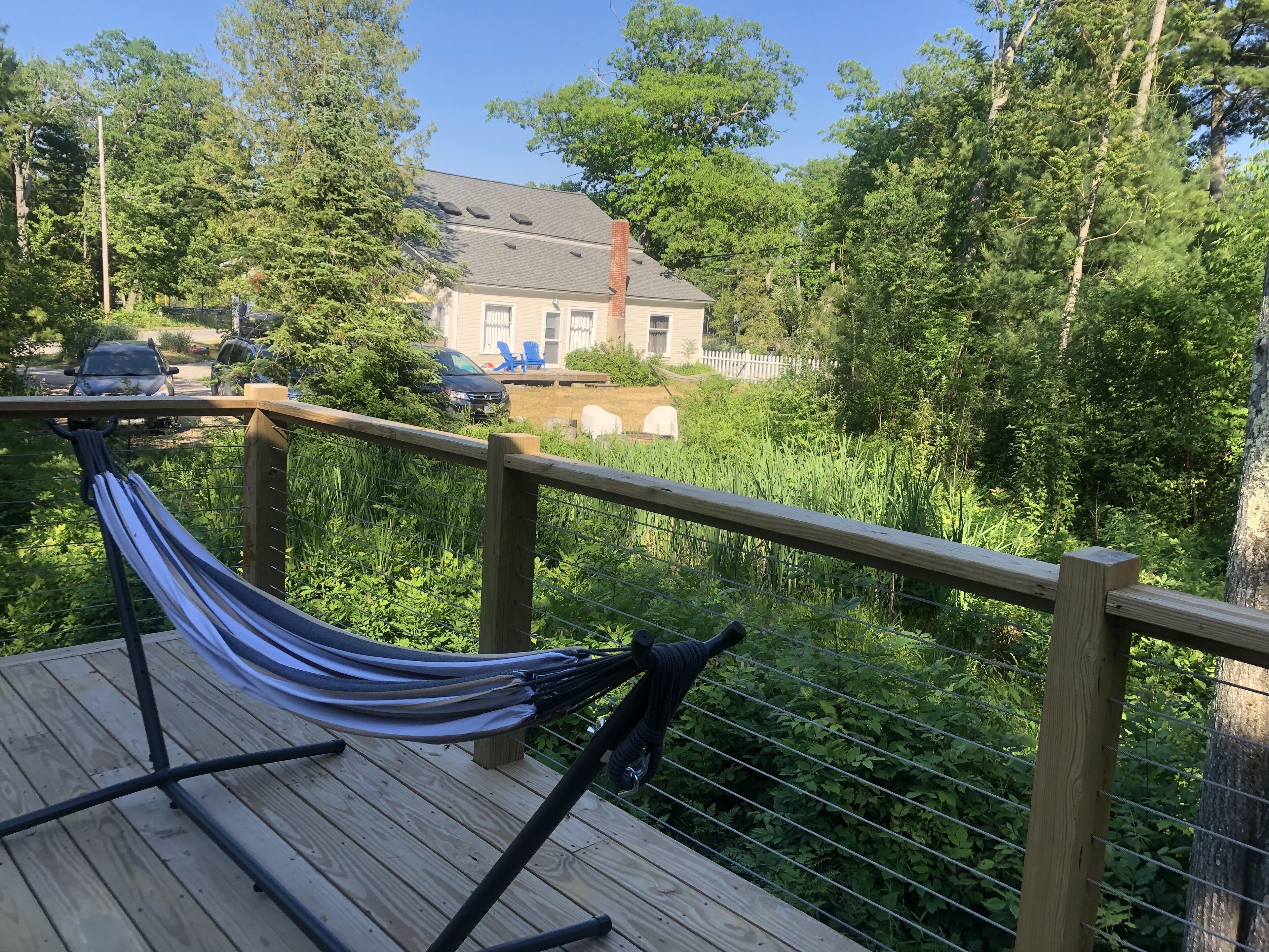 Una hamaca en una terraza de madera con vista a una casa y follaje circundante.