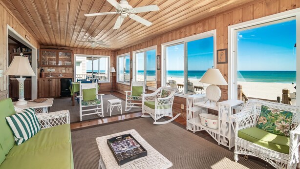 Amplia habitación con paredes y techo de madera, ventanas amplias con vistas a la playa, zona de comedor con mobiliario de mimbre blanco y una cocina visible a través de una puerta abierta.