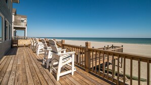 Terraza de madera con sillas blancas y barandilla de madera con vista a una playa.