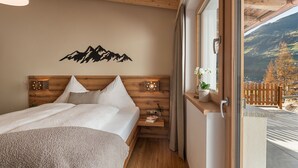 A bedroom with a wooden headboard, a bed with white bedding, a nightstand with a lamp, and a view of a mountainous landscape through a large window.