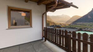A balcony with a wooden railing, a window with a light fixture, and a view of a valley with mountains in the background.