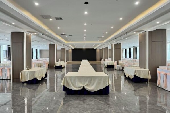 Une vaste salle de banquet avec une longue table au centre, entourée de tables plus petites recouvertes de nappes blanches et agrémentées de rubans roses.
