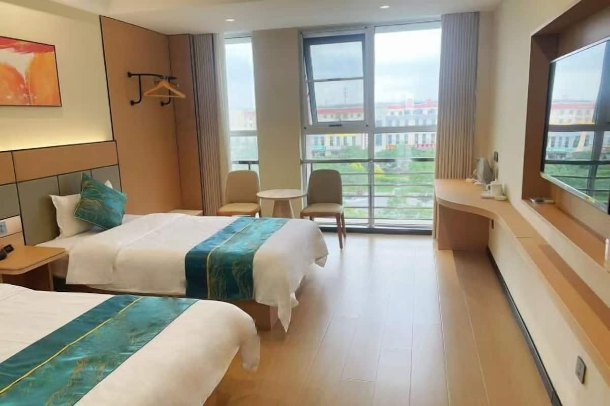 A hotel room with two beds, a small table, a chair, a large window, and a view of buildings outside.