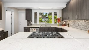 Una cocina moderna con armarios oscuros, una isla grande con estufa a gas, un ventanal con vistas a zonas verdes y un respaldo de azulejos.
