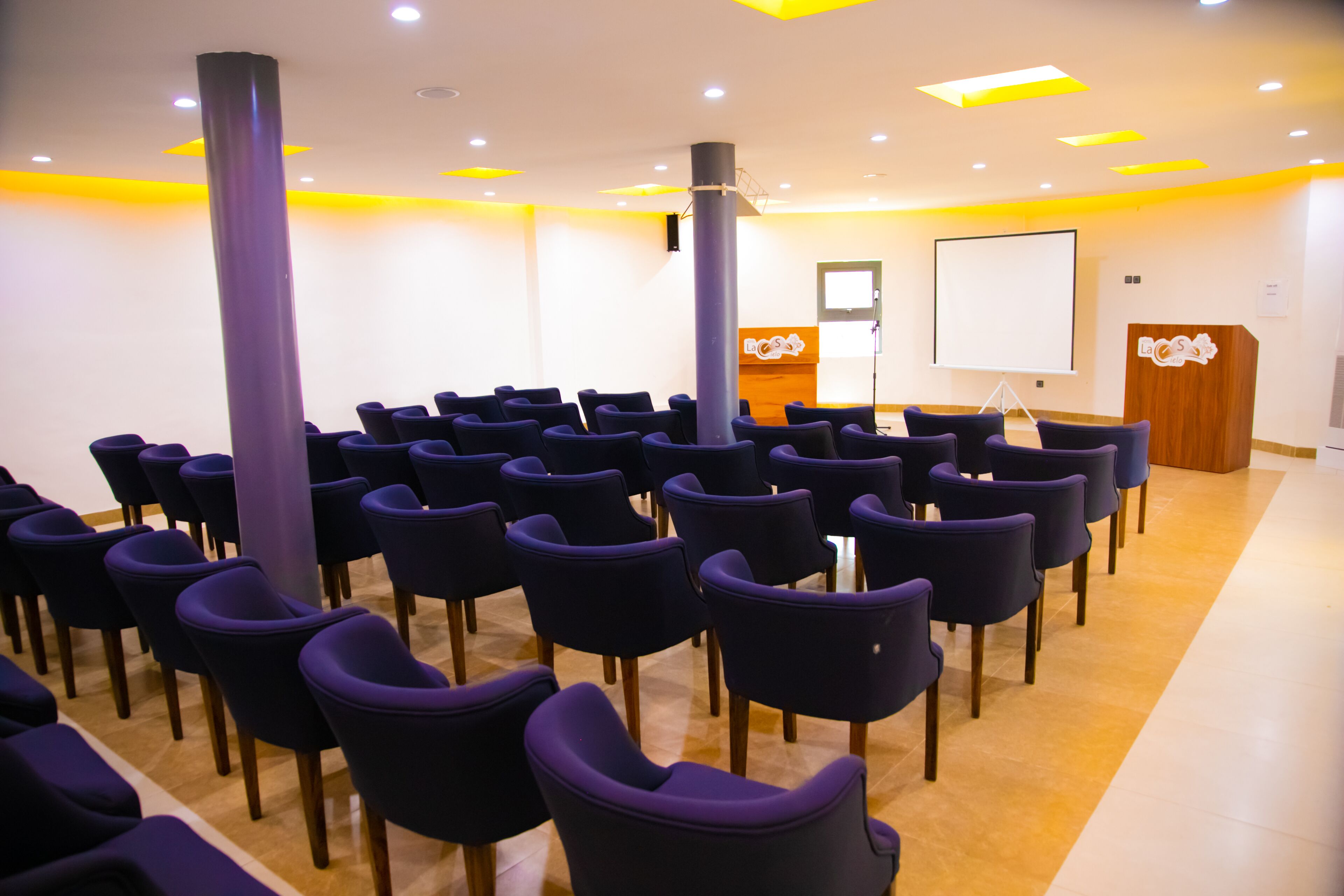 Un salón de conferencias con filas de sillas, un atril, una pantalla y un mostrador de recepción.