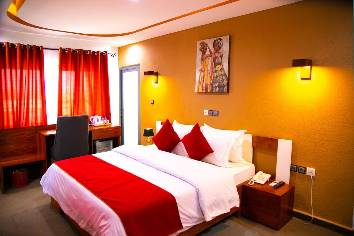 A hotel room with a large bed, red and white bedding, a desk with a chair, a telephone, and a painting on the wall.