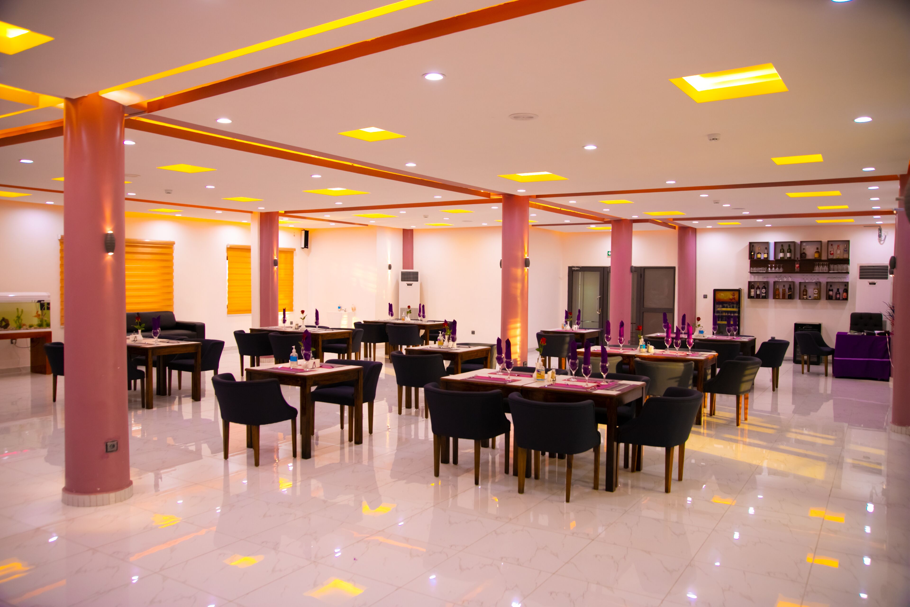 A spacious hotel lobby with multiple dining tables and chairs, a reception area, and a bar with framed pictures on the wall.