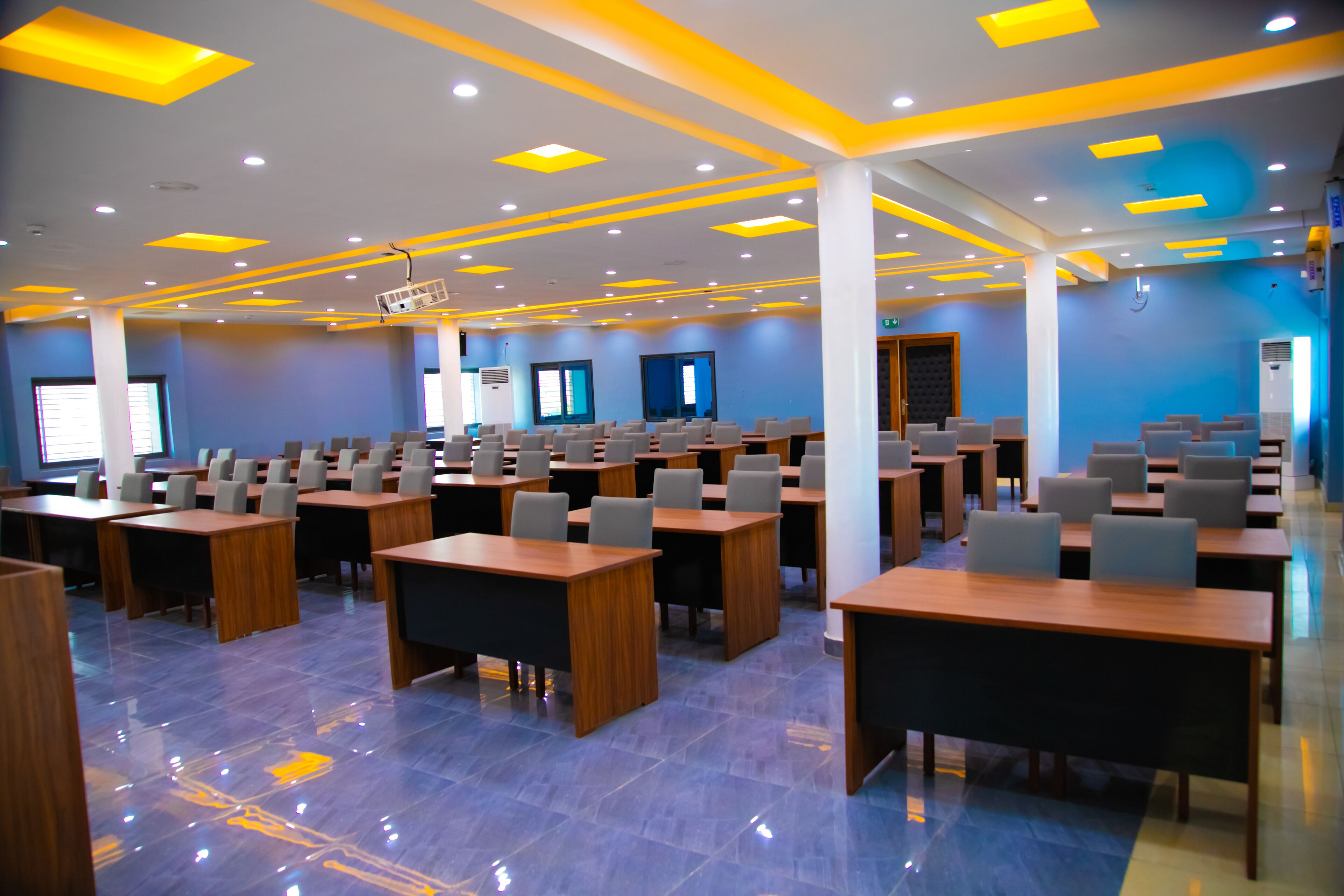 Un salón de conferencias con varias mesas y sillas, una pantalla de proyector y una pared azul con ventanas.