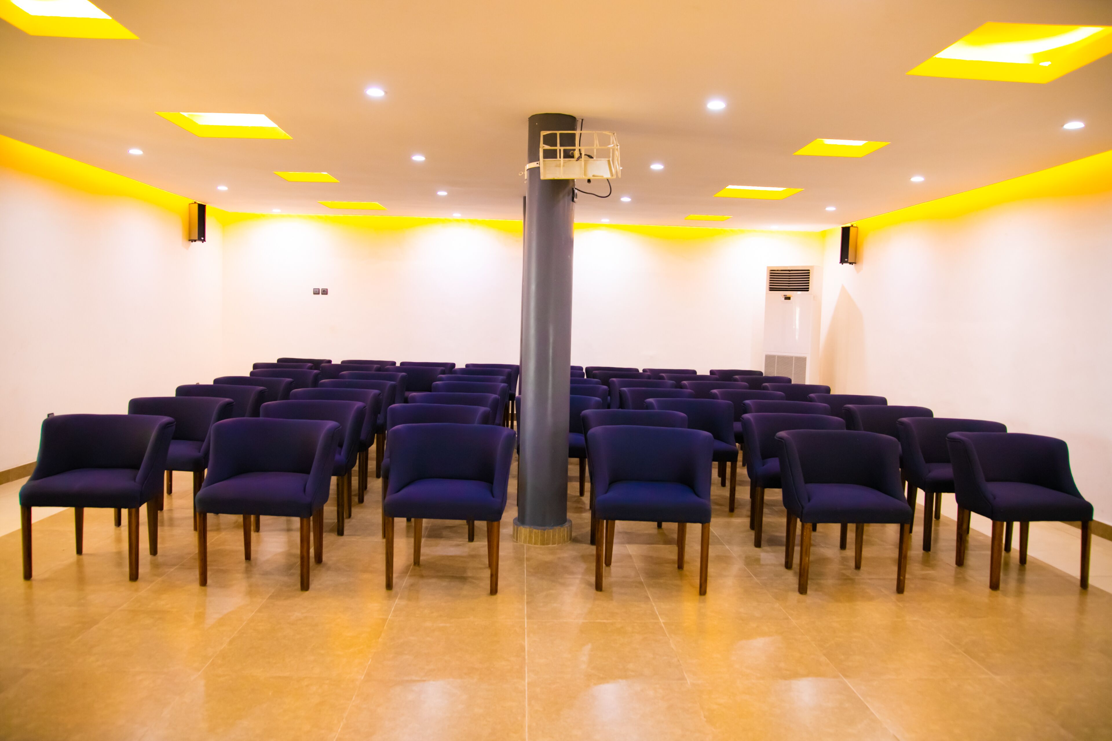 A conference room with rows of purple chairs, a central pillar, and yellow ceiling lights.