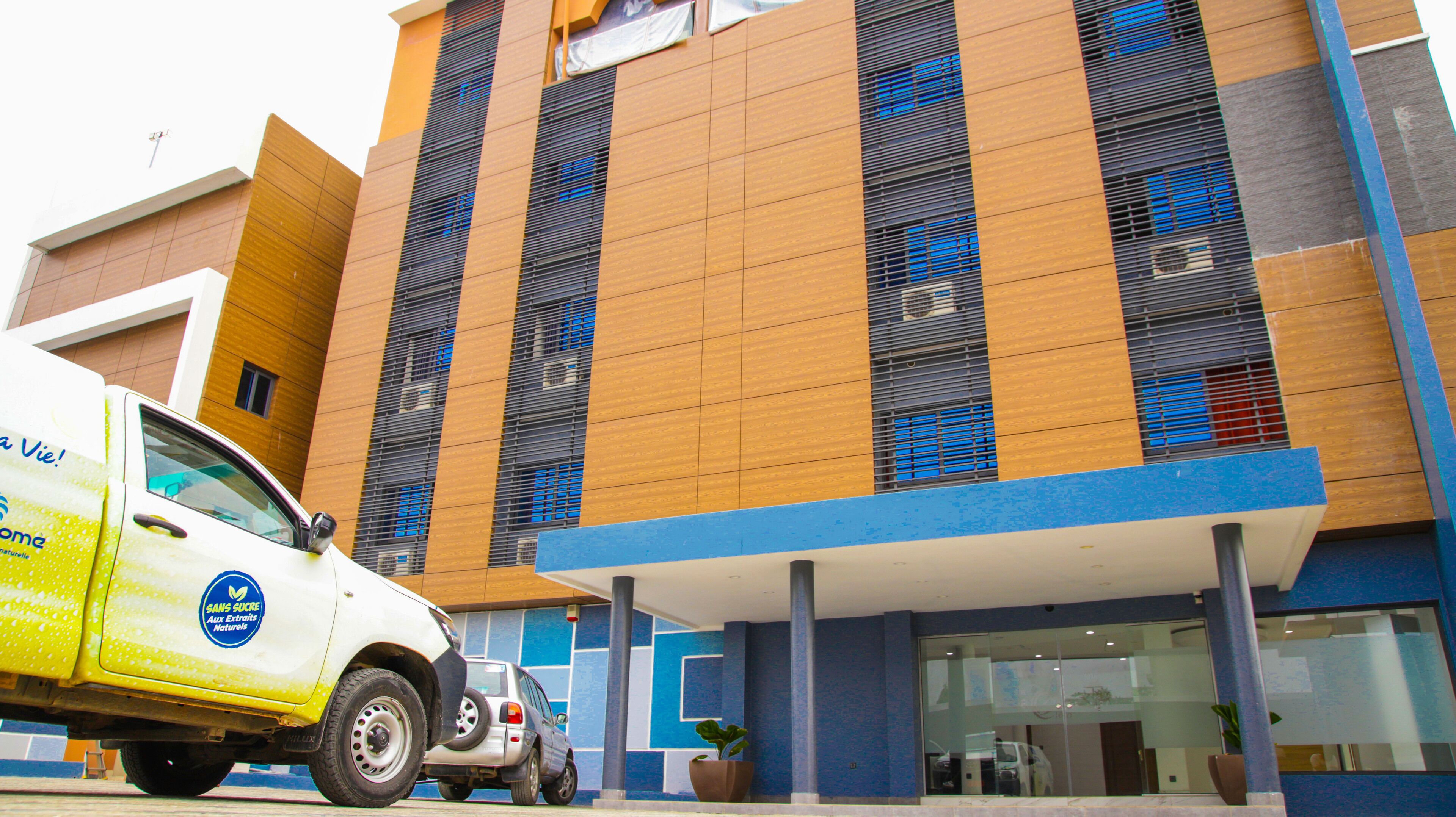 A modern building with a blue and brown facade, a white van parked in front, and a potted plant near the entrance.