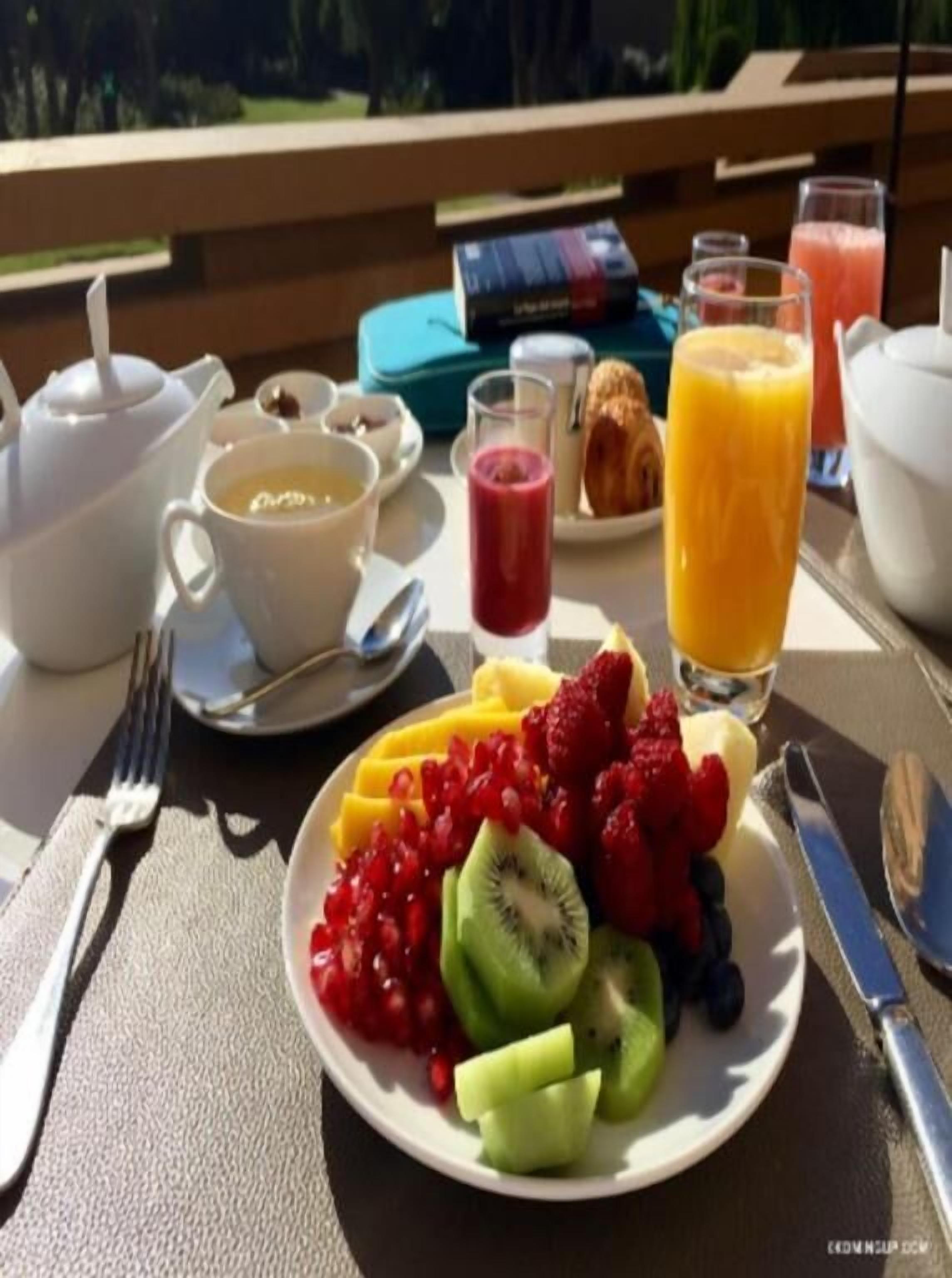 Een ontbijtsetting met een fruitschaal, een theepot, kopjes en een glas sinaasappelsap op een tafel.