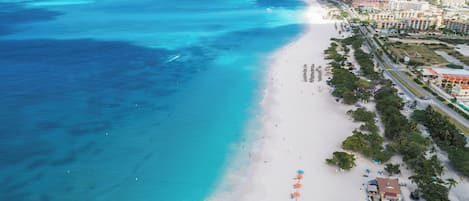 A coastal beach area with a white sandy shore, clear turquoise waters, and a row of buildings along the beachfront.