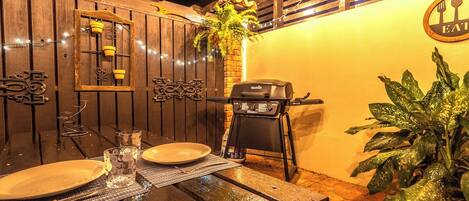 Een buitenterras met een houten tafel, stoelen, een barbecue en een plant in pot.
