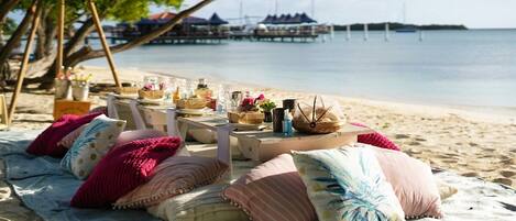 Ein Essbereich am Strand mit Tischen, Kissen und Blick auf das Meer und den Steg.