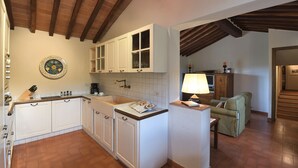 A kitchen with white cabinets, a tiled backsplash, and a wooden ceiling. There is a dining area with a green sofa and a lamp. - Dionisia, Capannole, Gaiole in Chianti, Siena and Chianti (Gaiole in Chianti)