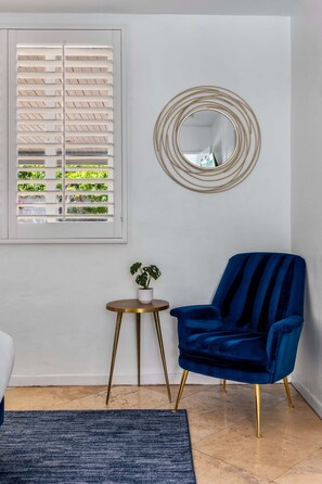 A room with a window with blinds, a round mirror, a blue chair, a small table with a plant, and a blue rug. - Thanks for the Memory – Bob Hope Historic Estate (Palm Springs)