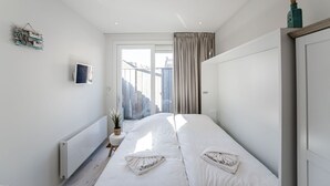 Ein modernes Schlafzimmer mit einem großen Bett, einem Heizkörper, einem kleinen Tisch mit Pflanze und Blick ins Freie durch eine Glastür.