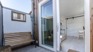 Ein modernes Interieur mit einer Holzbank, einer Glastür zu einem Raum mit Bett und Sessel sowie einem kleinen Tisch mit Pflanze.