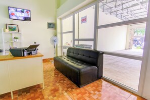 A reception area with a leather sofa, a desk with a laptop, a TV displaying multiple screens, and a large window with a 'OYO' sign. - OYO 3493 Nami House (Medan)