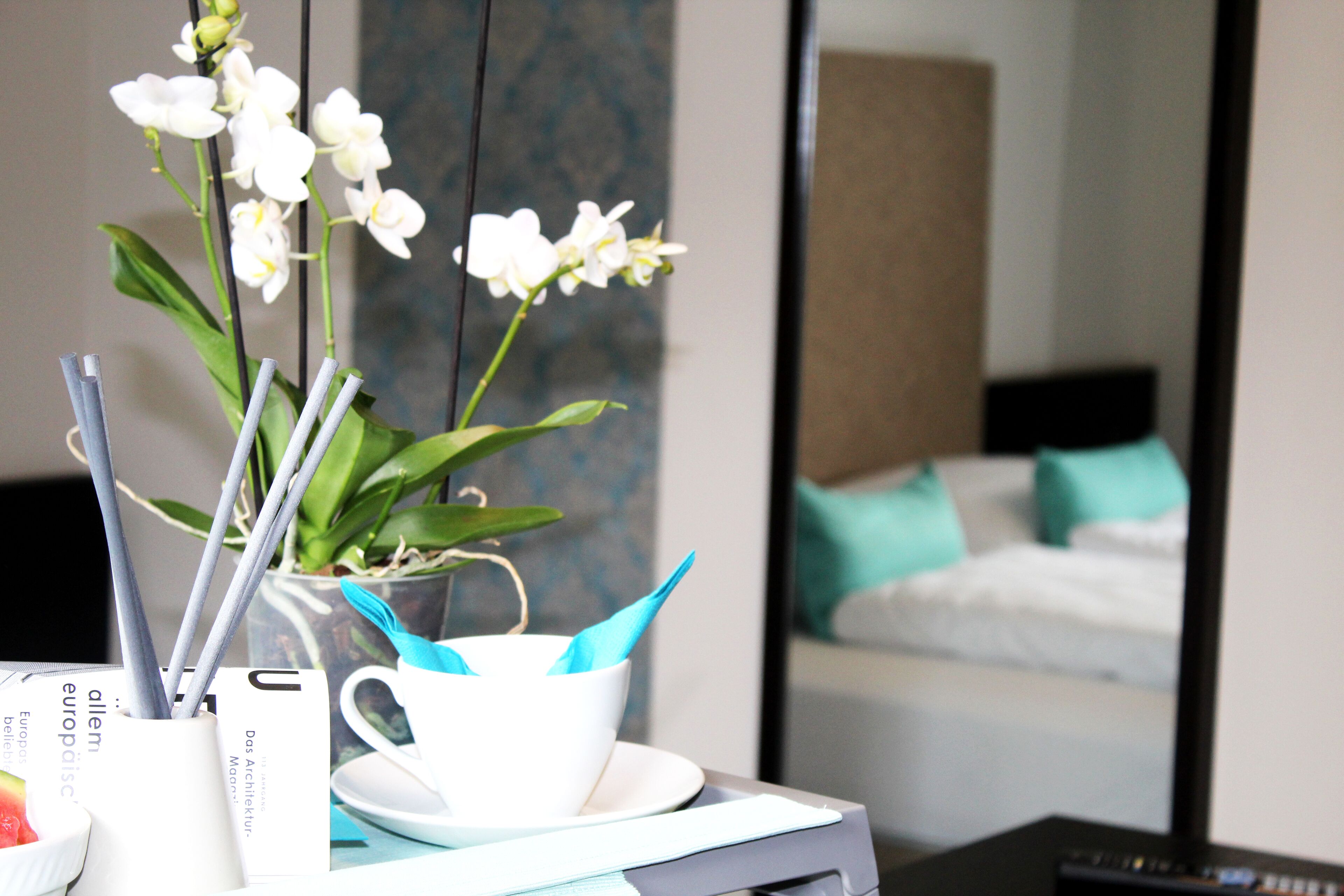 Habitación de hotel con cama, un espejo, una planta de orquídea blanca y una taza blanca con servilleta azul.