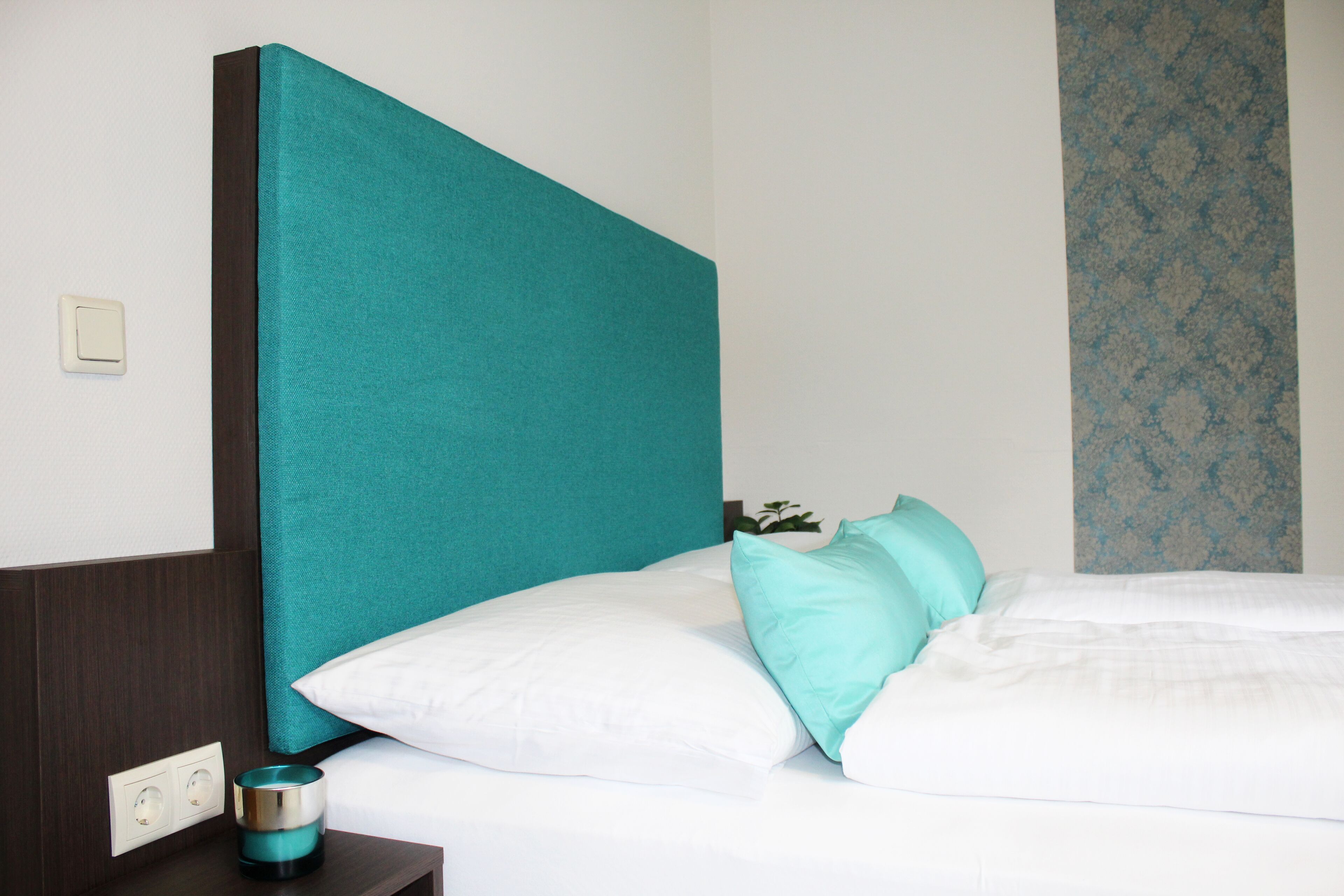 A neatly made bed with white and teal bedding, a teal headboard, and a small bedside table with a teal candle.