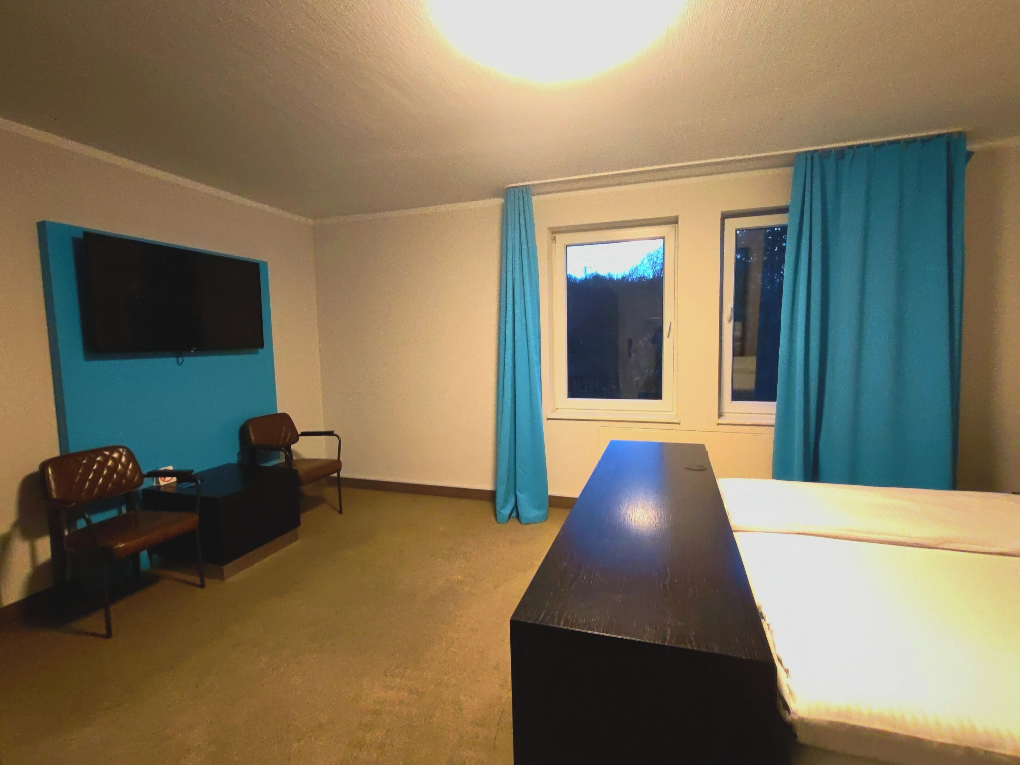 A room with a bed, a desk, a chair, a TV, and two windows with curtains.