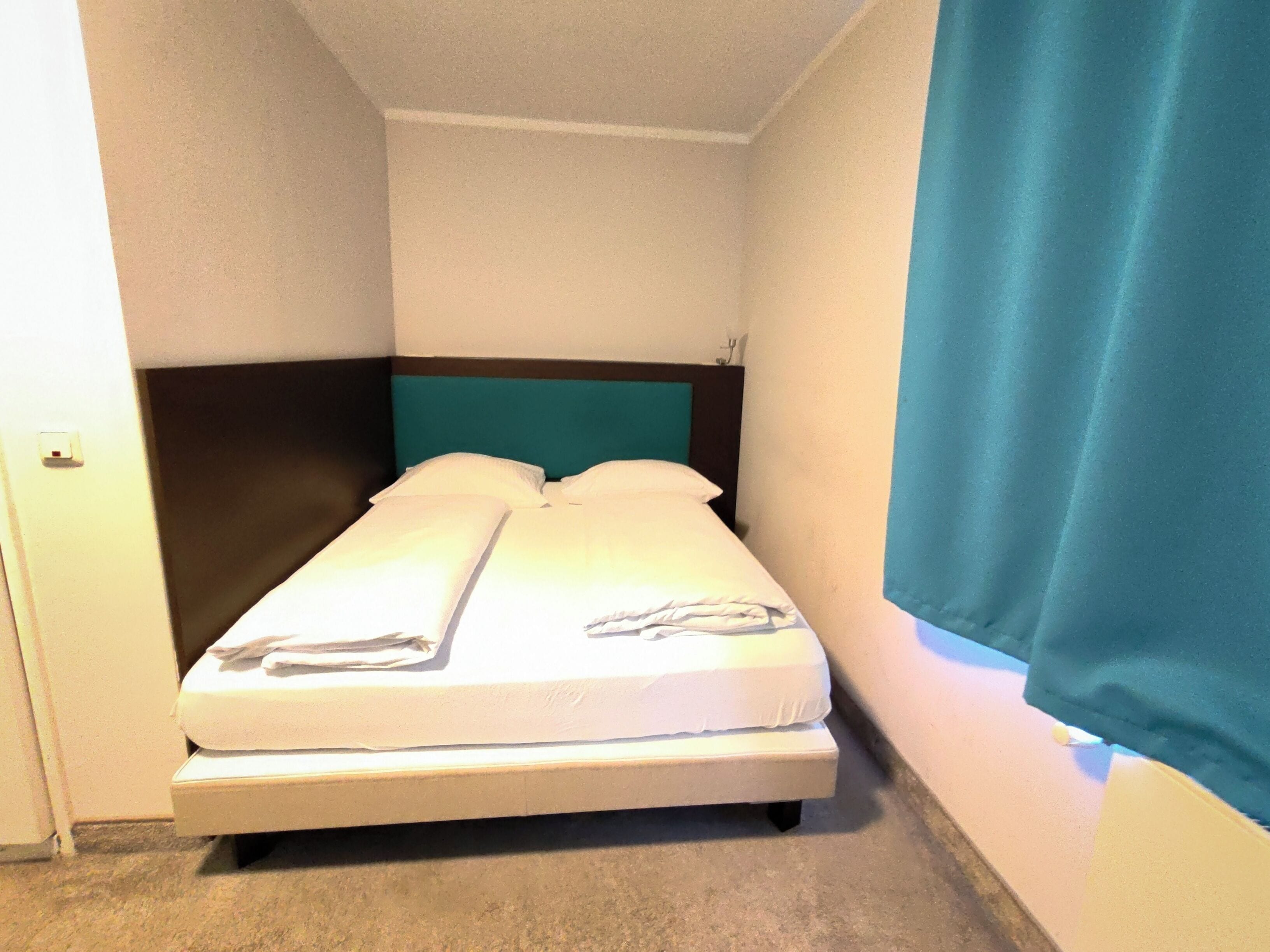 Ein kleines Zimmer mit einem Einzelbett, einem türkisblauen Kopfteil, weißer Bettwäsche und einer schlichten Wand.