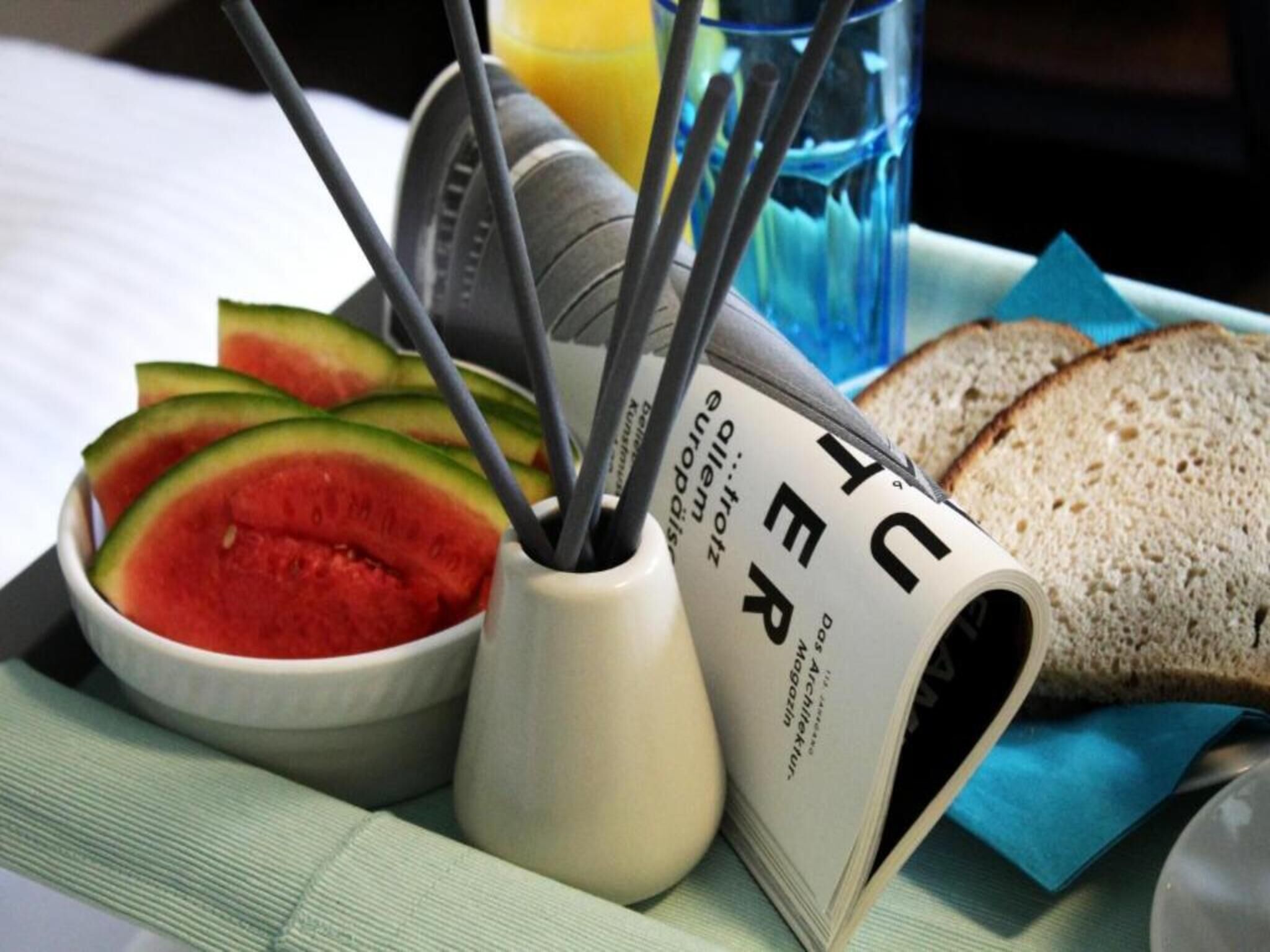 A breakfast tray with sliced watermelon, a glass of orange juice, a book, and a slice of bread.