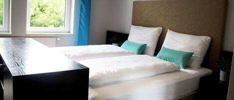 A double bed with white and teal pillows, a wooden headboard, and a window with a view of trees.