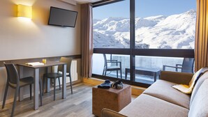 A modern hotel room with a large window offering a mountain view, a dining area with a table and chairs, and a cozy seating area with a sofa and ottoman. - SOWELL RÉSIDENCES Crêt Voland (Les Belleville)