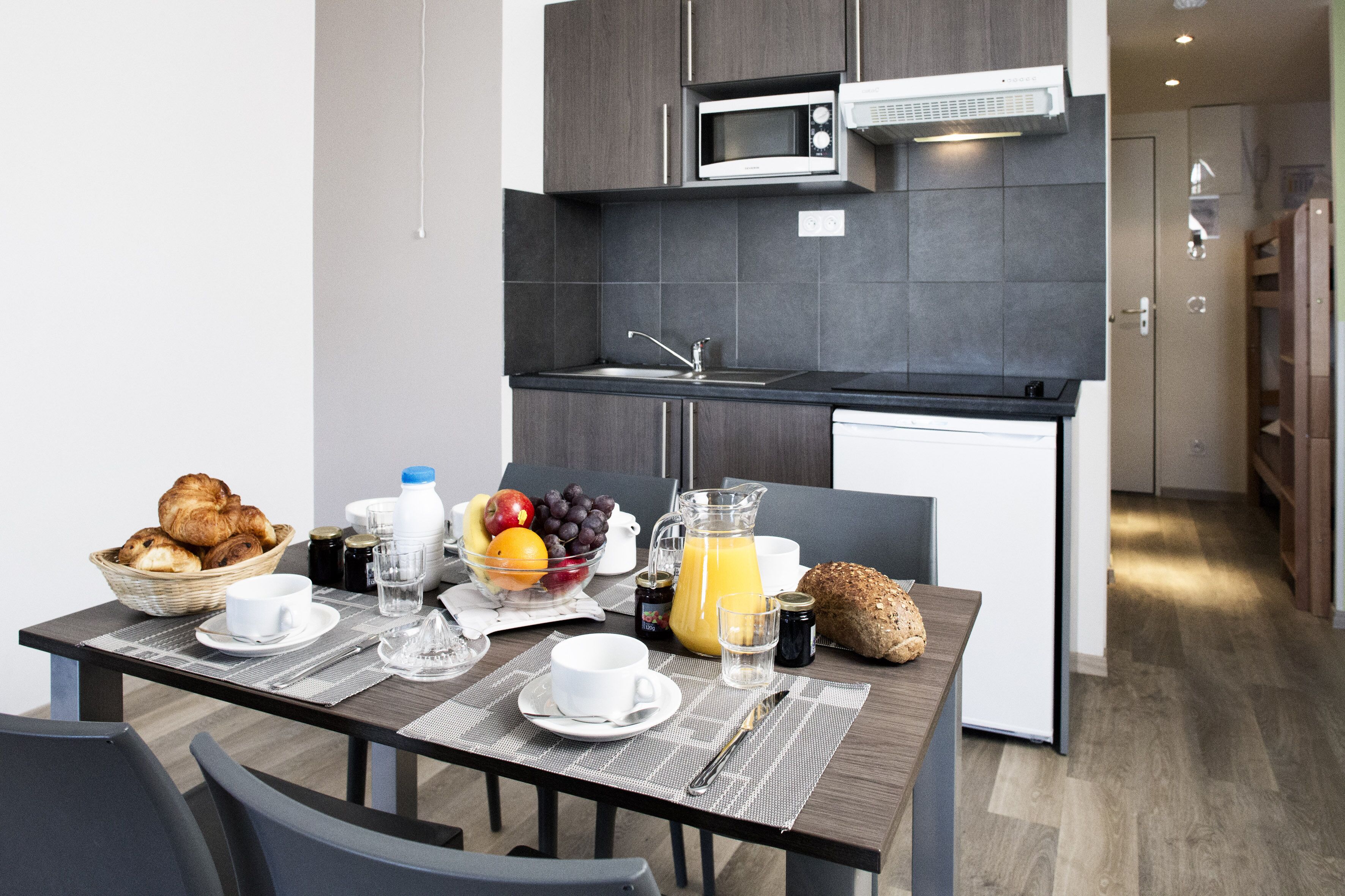 Eine moderne Küche mit dunklen Schränken, einer Mikrowelle und einem Spülbecken. Ein Esstisch ist gedeckt mit Croissants, Obst, Saft und Kaffee.