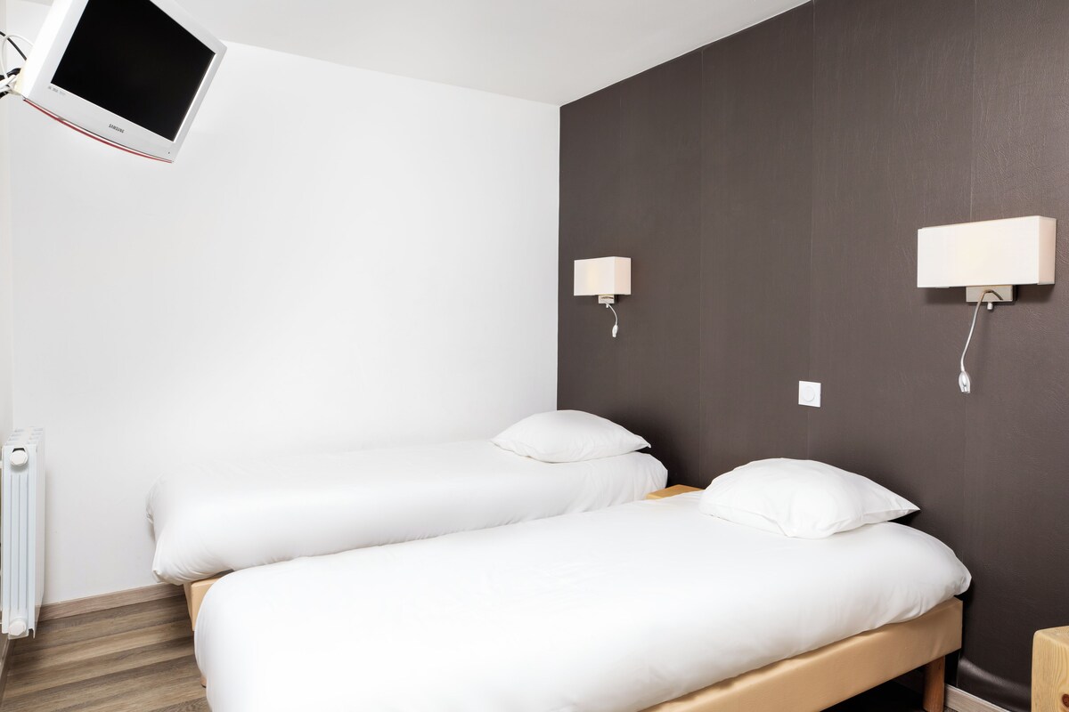 A hotel room with a bed, a TV mounted on the wall, and two wall-mounted lamps.