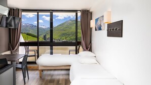 A modern hotel room with a large window offering a mountain view, a bed, a desk, and a chair. - SOWELL RÉSIDENCES Crêt Voland (Les Belleville)