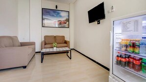 A living room with a beige sofa, a brown armchair, a coffee table with a vase, a flat-screen TV mounted on the wall, and a refrigerator stocked with beverages.