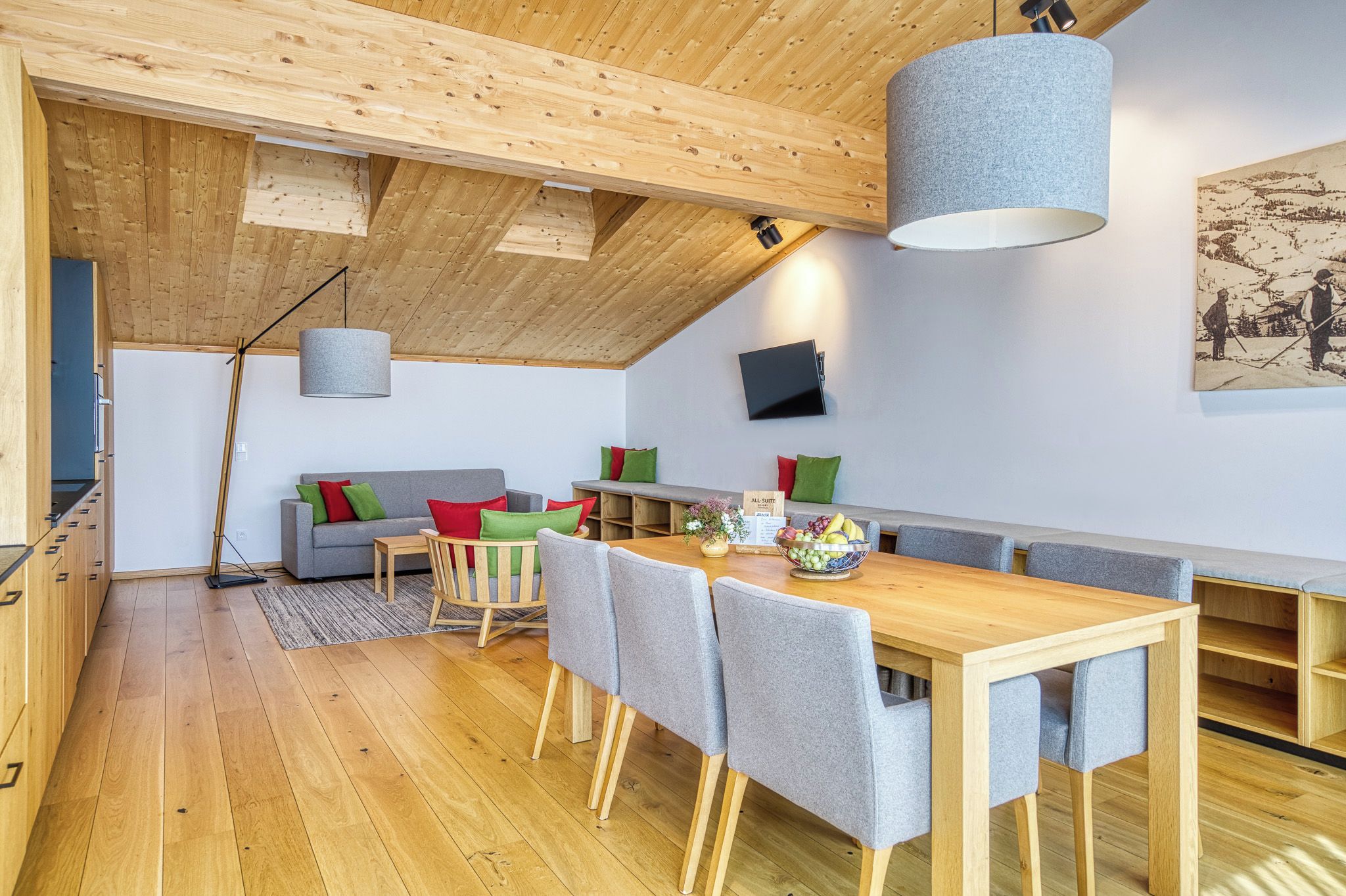A modern interior with a wooden ceiling, a dining area with a table and chairs, a sofa with cushions, and a mounted television.