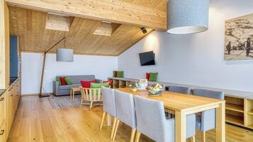 A modern interior with a wooden ceiling, a dining area with a table and chairs, a sofa with cushions, and a mounted television.