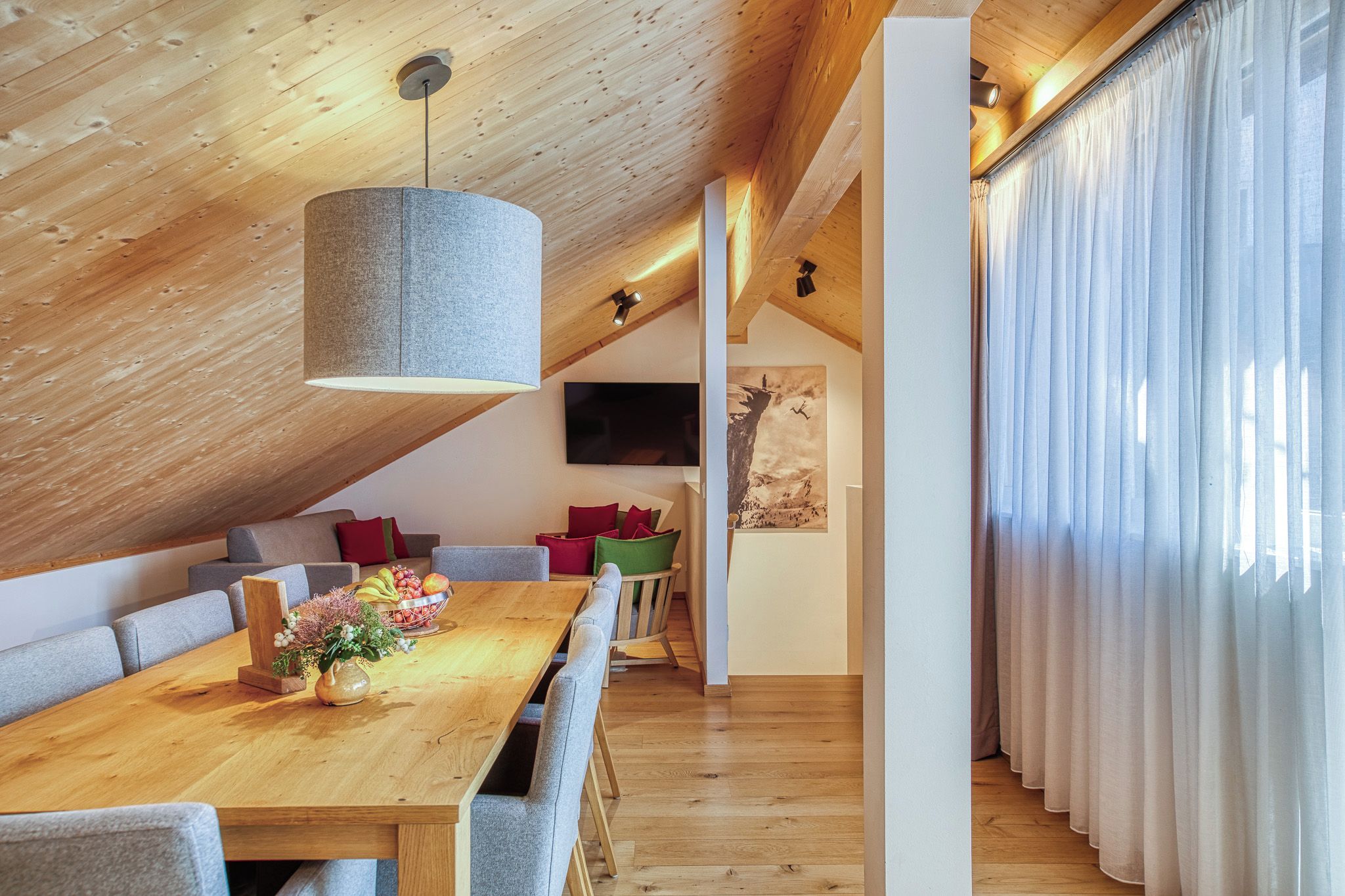 A modern loft-style room with a wooden ceiling, a dining area with a long table, and a large window with curtains.