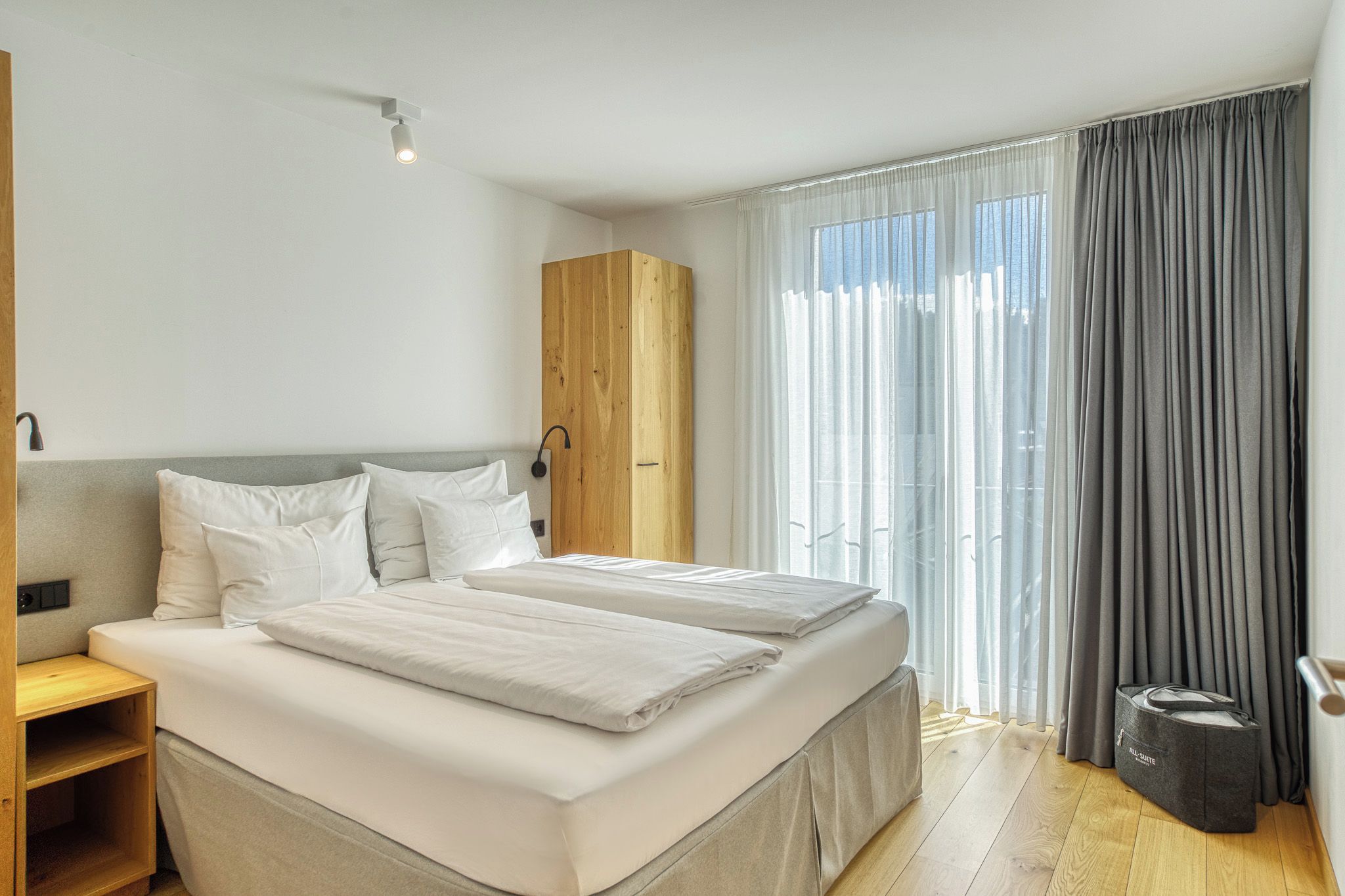 A modern hotel room with a double bed, a wooden wardrobe, a bedside table, and a large window with curtains.