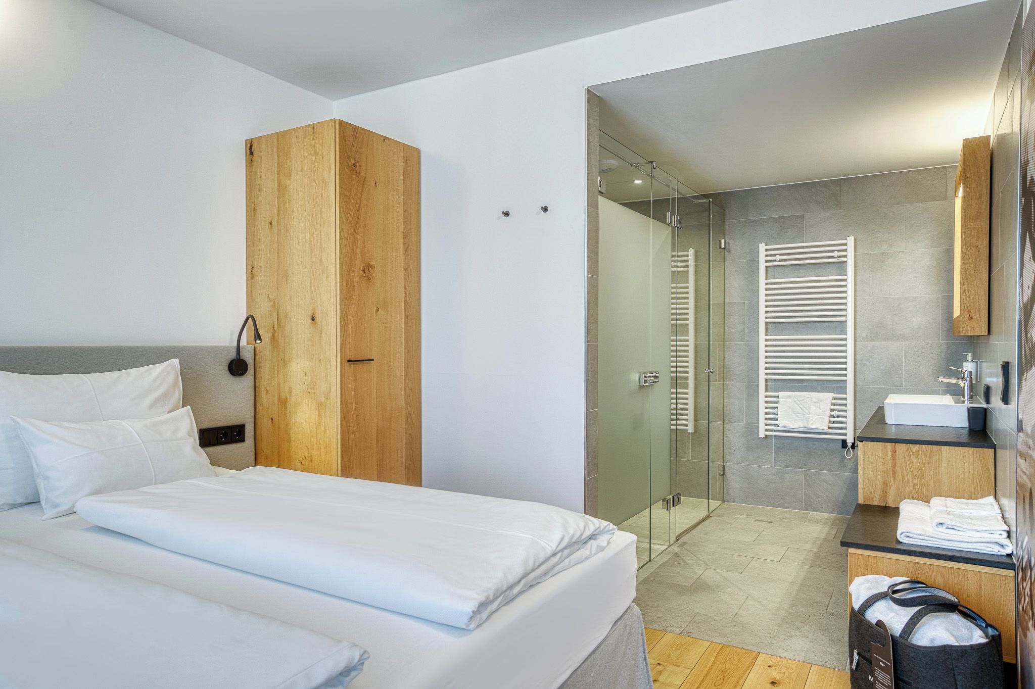 A modern hotel room with a bed, a wardrobe, a shower area, and a bathroom with a sink and towel rack.