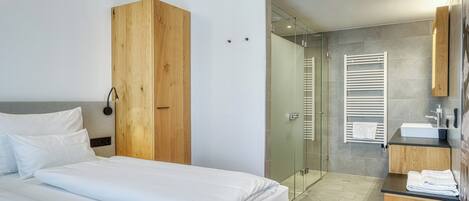 A modern hotel room with a bed, a wardrobe, a shower area, and a bathroom with a sink and towel rack.