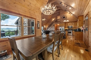 A wooden cabin interior with a dining area, kitchen, and a large window overlooking a landscape. - Fire on the Mountain - Mountain Views | 3 Levels of Decks | Outdoor Fireplaces | Hot Tub | Game Room (Mineral Bluff)