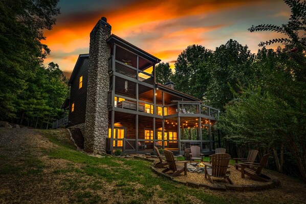 A two-story cabin with a stone chimney, large windows, and a deck area with outdoor seating.